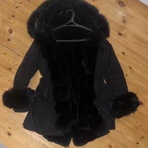 Svart parkajacka med pälsdetaljer - Snygg svart parkajacka med fluffiga pälsdetaljer på huva och ärmslut. Perfekt för kyliga dagar med sin stilrena design och varma känsla. Jackan har en dragkedja framtill och praktiska fickor. 