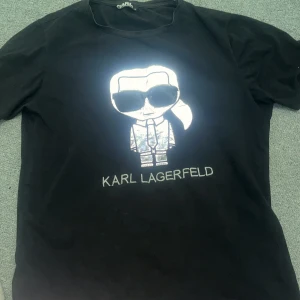 Svart t-shirt med tryck från Karl Lagerfeld - Snygg svart t-shirt från Karl Lagerfeld med ett coolt tryck av en figur på framsidan. Perfekt för dig som vill ha en stilren look med en twist. T-shirten är kortärmad och gjord i ett bekvämt material.