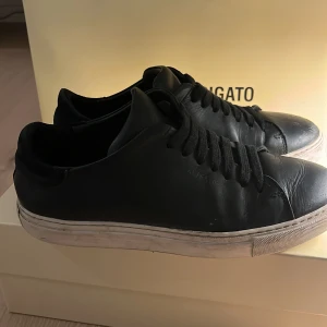 Svarta sneakers från Axel Arigato - Snygga svarta sneakers från Axel Arigato med klassisk design och snörning. Skorna har en vit sula och är tillverkade i skinn, vilket ger en stilren look. Perfekta för en trendig vardagsstil. Skorna är köpta på NK för cirka 2500kr. Priset kan diskuteras.