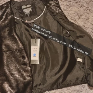 Svart paljettväst - Snygg svart väst täckt med glittriga paljetter. Perfekt för att lägga till lite glamour till din outfit. Västen har en öppen design och är idealisk för festliga tillfällen.