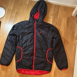 Svart quiltad jacka från Sweet Protection - Säljer en svart quiltad jacka från Sweet Protection med röda detaljer. Jackan har en huva och dragkedja framtill samt två fickor med röda kanter. Perfekt för kyligare dagar eller passar utmärkt under skaljackan.