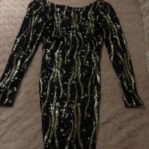 Kort svartguld klänning - Snygg svart klänning med guldiga paljetter i ett vågigt mönster. Klänningen har långa ärmar och en figurnära passform, perfekt för en festlig kväll som t.ex nyår eller Julafton.. Den glittriga designen ger en lyxig känsla. Den är från Soky & soka, pris kan diskuteras 😁