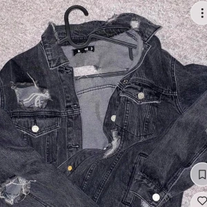 Svart jeansjacka med slitningar - Cool svart jeansjacka med slitna detaljer och knappar framtill. Jackan har en klassisk krage och två bröstfickor med lock. Perfekt för en avslappnad stil.