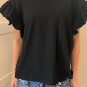 Svart topp med volangärmar - Snygg svart topp med volangdetaljer på ärmarna. Perfekt för en stilren look. Passar bra till jeans eller kjol.