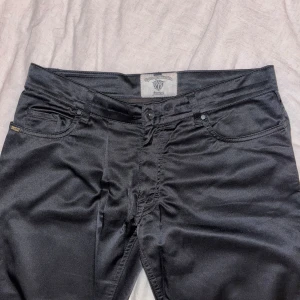 Svarta byxor från Tiger of Sweden - Snygga svarta byxor från Tiger of Sweden med en stilren design. Byxorna har en normal passform och är tillverkade i ett glansigt material som ger en lyxig känsla. Perfekta för en trendig look.