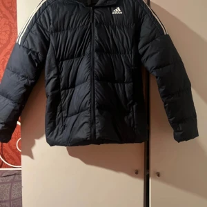 Svart dunjacka från Adidas - Snygg svart dunjacka från Adidas med vita ränder på ärmarna. Jackan har en dragkedja framtill och är perfekt för kyligare väder. Den har en sportig look med långärmade ärmar och en bekväm passform.användt bara 1 eller 2 gånger 