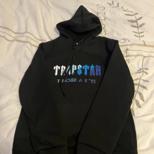 Svart hoodie och byxor från Trapstar - Säljer en svart hoodie och matchande byxor från Trapstar. Hoodien har en stor ficka framtill och tryck i blått och vitt på bröstet. Byxorna har en bekväm passform med resår och snörning i midjan samt tryck på sidan. Perfekt för en avslappnad stil.