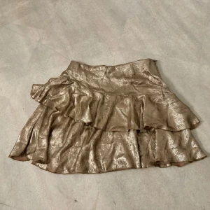 Glittrig guld kjol med volang - Snygg glittrig guld kjol med volangdetaljer. Perfekt för att sticka ut med en glamorös look. Kjolen har en kort längd och är tillverkad i ett skimrande material som fångar ljuset fint.