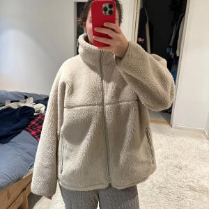 Beige teddyjacka med dragkedja - Mysig beige teddyjacka med dragkedja och hög krage. Perfekt för kyliga dagar med sin fluffiga och varma design. Jackan har långa ärmar och en avslappnad passform. Nypris 1250 kr.