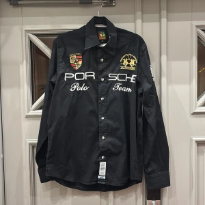 Svart skjorta från La Martina - Snygg svart skjorta från La Martina med broderade detaljer och texten 'Porsche Polo Team'. Skjortan har långa ärmar och knappar framtill. Perfekt för den stilmedvetna.