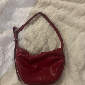 Röd axelväska med dragkedja - Snygg röd axelväska i glansigt material med justerbar axelrem och dragkedja upptill. Perfekt för att bära dina nödvändigheter med stil.