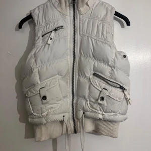 Vit dunväst från Vintage Edition - Säljer en snygg vit dunväst från Vintage Edition med dragkedja och flera praktiska fickor. Västen har en ribbad krage och nederkant för extra komfort. Perfekt för kyligare dagar!