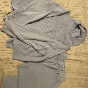 Blå/Grå ribbad hoodie & ribbade byxor - Säljer en stilren blå/grå ribbad hoodie med långa ärmar och blå/grå ribbade byxor till. Perfekt för en avslappnad look. Den har en bekväm passform och är gjord i ett mjukt material. Passar från:42-48