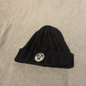 Svart stickad mössa från North Sails - Snygg svart stickad mössa från North Sails med ett broderat märke framtill. Perfekt för kyliga dagar med sin ribbade kant som ger extra värme och stil.