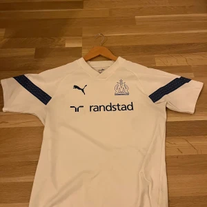 Vit fotbollströja från Puma - Säljer en vit fotbollströja från Puma med korta ärmar och mörkblå detaljer. Tröjan har en V-ringning och klubbmärke på bröstet. Perfekt för fotbollsfans!