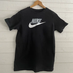 Svart barn t-shirt från Nike  - Snygg svart t-shirt från Nike med det klassiska logotyptrycket i vitt på bröstet. Perfekt för en sportig look eller avslappnad stil. Storlek 158-170cm/XL barn.