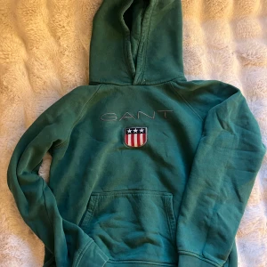 Grön hoodie från GANT - Säljer en grön hoodie från GANT med en broderad logga och ett emblem på bröstet. Tröjan har en klassisk känguruficka och en bekväm huva. Perfekt för en avslappnad stil.