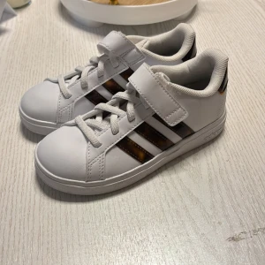 Vita sneakers från Adidas - Säljer ett par vita sneakers från Adidas med klassiska tre ränder i brunt på sidan. Skorna har snörning och kardborreband för extra säkerhet. Perfekta för en sportig look!