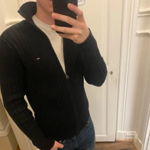 Svart ribbad kofta från Tommy Hilfiger - Snygg svart ribbad kofta från Tommy Hilfiger med dragkedja framtill. Den har en liten logotyp på bröstet och en hög krage. Perfekt för en stilren look. Modellen ca 182, tveka inte att kontakta vid eventuella frågor 😀