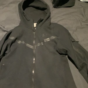 Svart nike tech fleece hoodie - Säljer en svart hoodie från Nike med dragkedja och huva. Den har en diskret logga på bröstet och stilrena detaljer i form av svarta band över bröstet och ärmarna. Perfekt för en casual look.