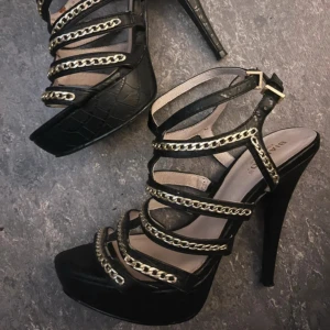 Svarta sandaler med kedjedetaljer - Eleganta svarta sandaler med hög klack och kedjedetaljer. Skorna har flera remmar med guldfärgade kedjor och en justerbar rem runt ankeln. Perfekta för en stilfull look.
