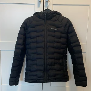 Svart dunjacka från Peak Performance - Säljer en svart dunjacka från Peak Performance. Jackan har en dragkedja framtill och är quiltad för extra värme. Perfekt för kyliga dagar med sin långa ärmar och huva. En stilren och funktionell jacka för vintern. (Har ett väldigt litet hål på sidan av armen på jackan och lite grön färg vid jackans ficka på vänstra sidan) jackan är otroligt fin och bra utöver det och väl värt pengarna, Nypris 1200kr! 