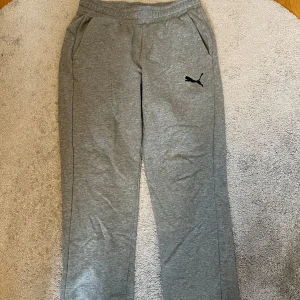 Grå mjukisbyxor från Puma - Säljer ett par grå mjukisbyxor från Puma med elastisk midja och sidofickor. Byxorna har en avslappnad passform och är perfekta för en bekväm dag hemma eller lätt träning. Puma-loggan är tryckt på ena benet.
