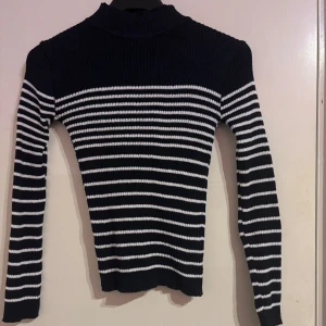 Svart och vit randig polotröja - Snygg svart och vit randig polotröja med ribbad design. Tröjan har långa ärmar och en tight passform, perfekt för kyligare dagar. Den klassiska randiga stilen gör den lätt att matcha med olika outfits.