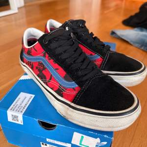 Snygga Vans sneakers med svart och röd färgkombination. Skorna har ett coolt mönster i rött och svart med blå detaljer på sidan. De har klassisk snörning och en vit sula som ger en skön kontrast.