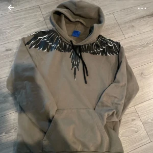 Grå hoodie från Marcelo Burlon - Säljer en grå hoodie från Marcelo Burlon med ett coolt vingmönster i svart och vitt runt halsen. Tröjan har en klassisk huva med svarta dragsnören och en stor magficka framtill. Perfekt för en stilren och avslappnad look.