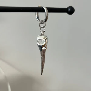 Silverfärgat örhänge i rostfritt stål med hängande detalj - Stilrent silverfärgat örhänge med en unik hängande fågel kranium. Perfekt för att ge en edgy touch till din stil. Örhänget har en slät yta och en enkel ringdesign.