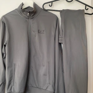 Set Emporio Armani - Säljer en stilren grå träningsjacka från Emporio Armani EA7. Jackan har en dragkedja framtill och en hög krage. Perfekt för träning eller en avslappnad dag. Den har en diskret logga på bröstet.