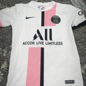  PSG fotbollströja - Säljer en vit PSG fotbollströja med rosa och svarta detaljer. Tröjan har korta ärmar och klubbens emblem på bröstet. Den har även sponsorloggor och messi på ryggen.
