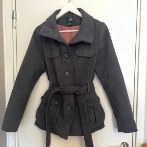 Mörkgrå kappa från H&M - Snygg mörkgrå kappa från H&M med knappar och bälte i midjan. Den har långa ärmar och två fickor framtill. Perfekt för kyligare dagar med sin stilrena design och bekväma passform.