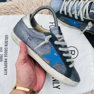 Glittriga sneakers från Golden Goose - Snygga glittriga sneakers från Golden Goose med en blå stjärna på sidan. Skorna har vita snören och en silverfärgad häl. Perfekta för att ge din outfit en extra touch av stil.