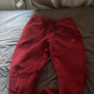 Vinröda nike tech byxor  - Säljer ett par bekväma röda mjukisbyxor från Nike med elastisk midja och snörning. Byxorna har en liten Nike-logga på framsidan och dragkedja på fickorna. Perfekta för en avslappnad stil.