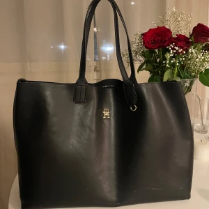 Svart shoppingväska från Tommy Hilfiger - Snygg och rymlig svart shoppingväska från Tommy Hilfiger i skinn. Väskan har ett elegant guldemblem på framsidan och en praktisk innerficka med dragkedja. Perfekt för att bära med sig allt man behöver under dagen. Finns några rivmärken från kattis, men fungerar väl. Hör av dig om du vill ha tydligare bilder 🌸Nypris: 1695kr 