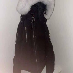 Svart dunjacka från Moose Knucklels - Säljer en svart dunjacka från Moose Knuckles med en lyxig vit päls på huvan. Jackan har dragkedja och flera snygga detaljer som metallknappar och broderad insida. Perfekt för kalla dagar, den kommer med påse för själva jackan och pälsen den är nästan helt ny använd 4-6 gånger den är i storlek xs enligt mig passar den S också , skriv om du funderar över något!