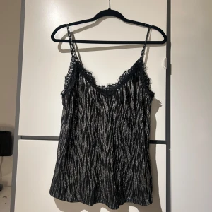 Svart glittrig linne med spetsdetaljer - Snygg svart linne med glittriga ränder och spetsdetaljer vid halsringningen. Tunna axelband och en lätt och luftig passform.