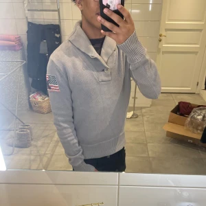 Grå stickad tröja från Polo Ralph louren  - Säljer en stilren grå stickad tröja från Polo med en amerikansk flagga på ärmen. Tröjan har en unik krage med knappar och ribbade muddar vid ärmslut och nederkant. Perfekt för kyliga dagar!