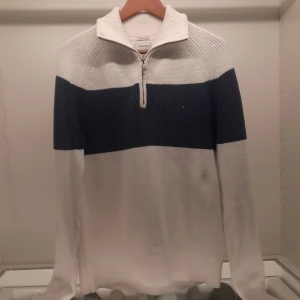 Vit och blå stickad tröja från Boomerang - Säljer en stilren vit och blå stickad tröja från Boomerang i 100% ekologisk bomull. Tröjan har en dragkedja vid halsen och en bred blå rand över bröstet. Perfekt för kyligare dagar med sin mjuka och bekväma passform.