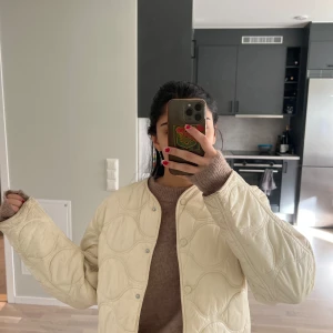 Täck jacka - Säljer en snygg beige quiltad jacka med rund hals och tryckknappar. Jackan har en stilren design och är perfekt för kyligare dagar. Den är lätt att matcha med olika outfits och ger en trendig look.