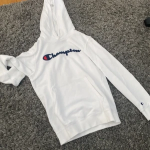 Vit hoodie från Champion - Säljer en snygg vit hoodie från Champion med deras ikoniska logga i blått och rött på bröstet. Tröjan har en stor ficka framtill och en bekväm huva. Perfekt för en avslappnad stil!