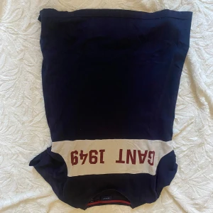 Mörkblå tröja från Gant - Säljer en snygg mörkblå tröja från Gant med beige och röd detalj. Tröjan har texten 'GANT 1949' tryckt över bröstet. Perfekt för en stilren och avslappnad look.
