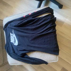 Svart långärmad tröja från Nike - Säljer en svart långärmad tröja från Nike med stort vitt logotryck på ryggen. Perfekt för en sportig look eller avslappnad stil. Tröjan är bekväm och passar till många tillfällen.