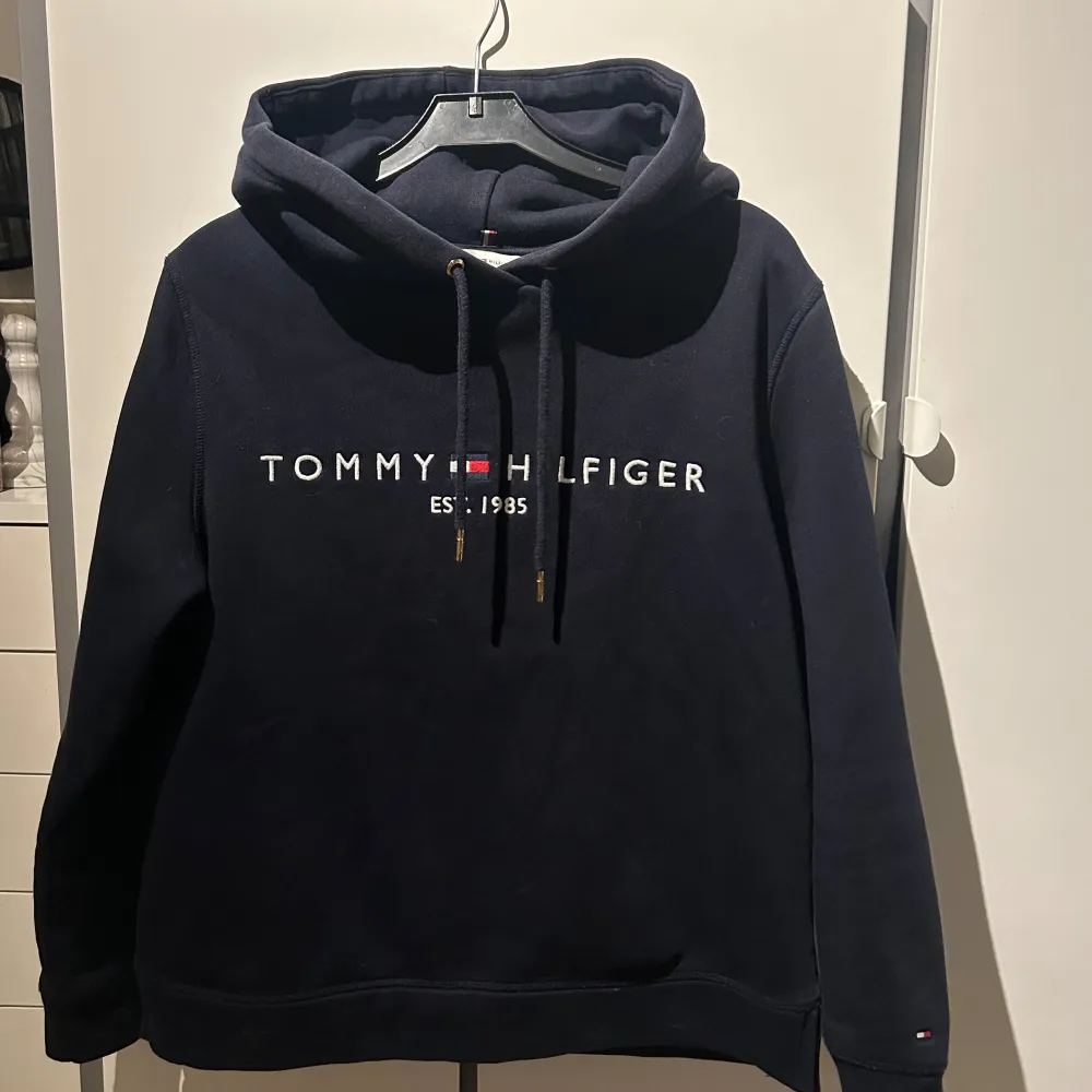 Säljer en snygg mörkblå hoodie från Tommy Hilfiger med logga och text framtill. Den har en klassisk design med justerbar huva och långa ärmar. . Neuletakit & Villapaidat.