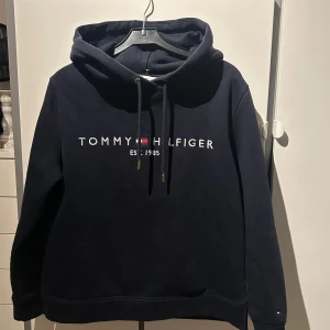 Mörkblå hoodie från Tommy Hilfiger - Säljer en snygg mörkblå hoodie från Tommy Hilfiger med logga och text framtill. Den har en klassisk design med justerbar huva och långa ärmar. 