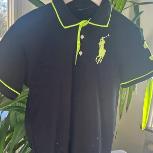 Svart pikétröja med gröna detaljer - Snygg svart pikétröja med neongröna detaljer från Polo. Tröjan har en broderad logga på bröstet och siffran 3 på ärmen. Perfekt för en cool look till sommaren. Sitter som en M, otroligt bra skick! Hör av dig vid minsta fundering.