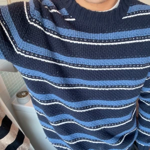 Randig stickad tröja från Mango - Säljer en snygg stickad tröja från H&M i blå och svart med vita ränder. Tröjan har en klassisk rund hals och långa ärmar, perfekt för kyligare dagar. Den är både stilren och bekväm, idealisk för en avslappnad look.