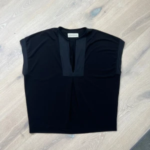 Svart topp från By Malene Birger - Snygg svart topp från By Malene Birger med en stilren design. Toppen har en djup v-ringning och är tillverkad i ett mjukt polyestermaterial med en liten del elastan för extra komfort. Perfekt för en elegant look.